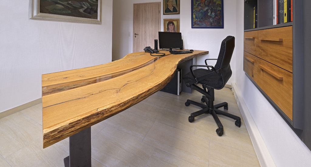 Massiver Schreibtisch aus heimischem Eichenholz mit Waldkante und maßgefertigtem Glaseinleger. Getragen von einem Stahlfuß links und einem Unterbaumöbel rechts.